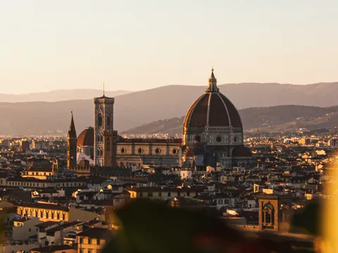 Vista panorámica de Florencia, Italia, al atardecer, destacando la Catedral de Santa María del Fiore con su cúpula de Brunelleschi y la torre del campanario, rodeadas de edificios históricos y montañas al fondo.
