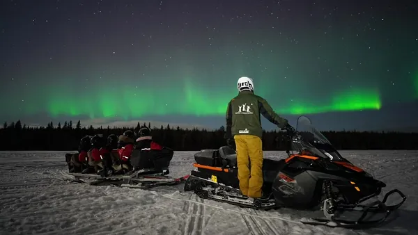 Motonieve negra y naranja con trineo sobre nieve, bajo auroras boreales verdes en el cielo nocturno.