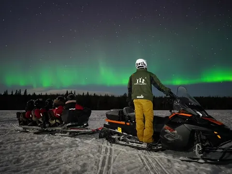 Motonieve negra y naranja con trineo sobre nieve, bajo auroras boreales verdes en el cielo nocturno.