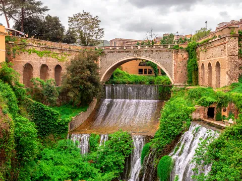 Cascada escalonada bajo puente histórico rodeado de vegetación y arquitectura tradicional.