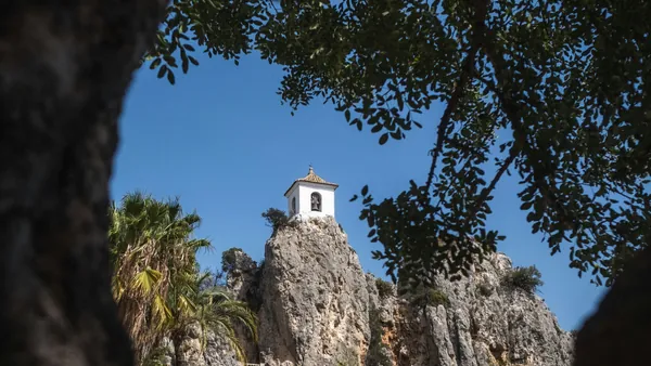 Campanario de Guadalest, un edificio blanco en la cima de una roca, visto a través de las ramas de los árboles.
