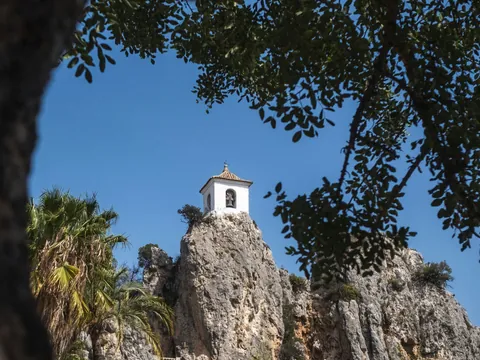 Campanario de Guadalest, un edificio blanco en la cima de una roca, visto a través de las ramas de los árboles.