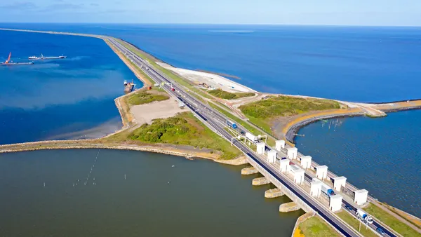 Vista aérea de una carretera y un dique (Afsluitdijk) que separa dos extensas masas de agua en los Países Bajos, con estructuras de control de agua y vehículos circulando, mostrando la ingeniería hidráulica holandesa.