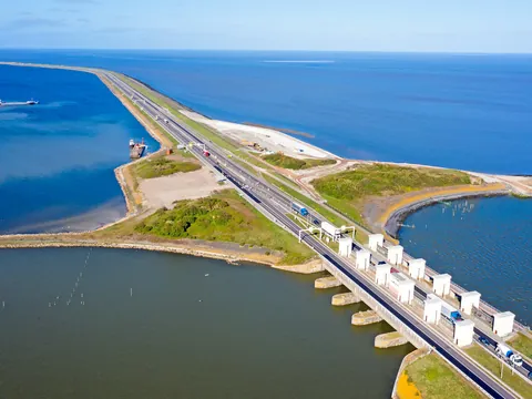 Vista aérea de una carretera y un dique (Afsluitdijk) que separa dos extensas masas de agua en los Países Bajos, con estructuras de control de agua y vehículos circulando, mostrando la ingeniería hidráulica holandesa.