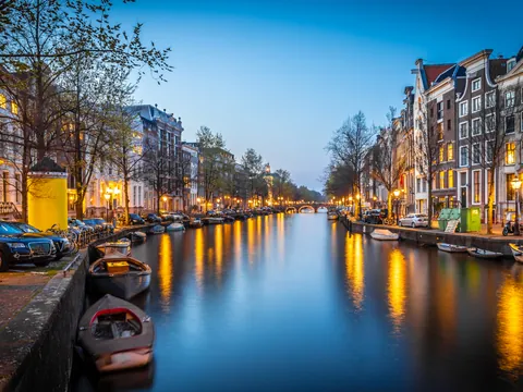 Canal de Ámsterdam al atardecer, bordeado por casas típicas neerlandesas, árboles y pequeñas embarcaciones, con luces reflejadas en el agua.