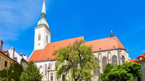 Catedral de San Martín en Bratislava, con su torre de aguja verde y tejado rojo, rodeada de árboles bajo un cielo azul claro.