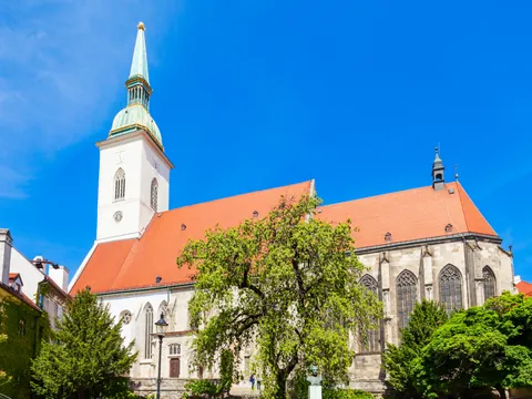 Catedral de San Martín en Bratislava, con su torre de aguja verde y tejado rojo, rodeada de árboles bajo un cielo azul claro.