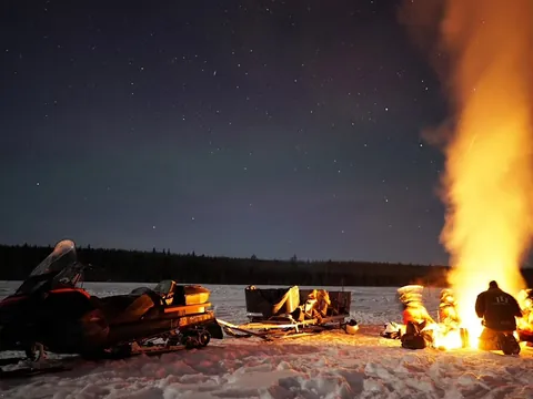 Grupo alrededor de una gran fogata en la nieve, junto a motonieve y trineos, bajo un cielo nocturno estrellado.