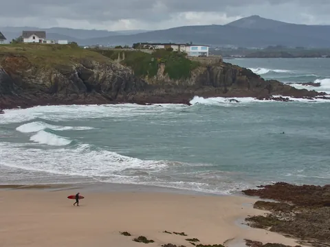 Surfista con tabla roja caminando por la playa de Tapia de Casariego, con el mar Cantábrico y acantilados al fondo.