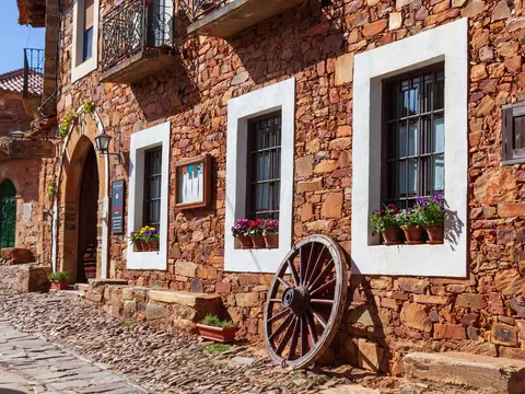 Calle de Castrillo de los Polvazares, León, con casa maragata de piedra rojiza, ventanas encaladas y rueda de carro.
