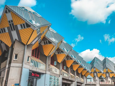 Fila de casas cúbicas amarillas y grises con techos inclinados en Rotterdam, conocidas por su diseño arquitectónico innovador, bajo un cielo azul brillante con algunas nubes blancas.