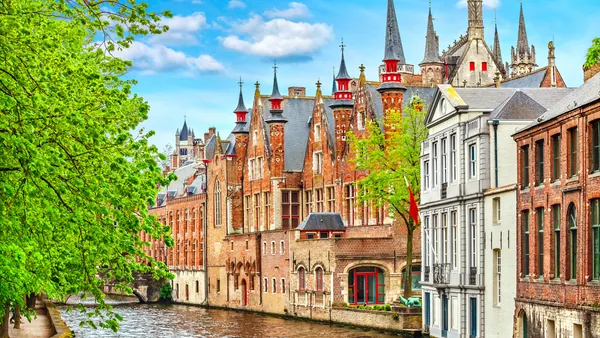 Canal de Brujas en Bélgica rodeado de edificios históricos de ladrillo, puentes y vegetación, con reflejos en el agua bajo un cielo parcialmente nublado.