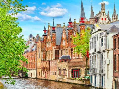 Canal de Brujas en Bélgica rodeado de edificios históricos de ladrillo, puentes y vegetación, con reflejos en el agua bajo un cielo parcialmente nublado.