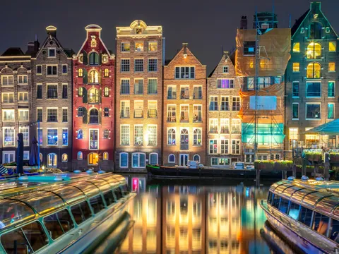 Fachada de casas estrechas y coloridas iluminadas por la noche, reflejadas en el canal de Ámsterdam, con barcos turísticos atracados en primer plano.