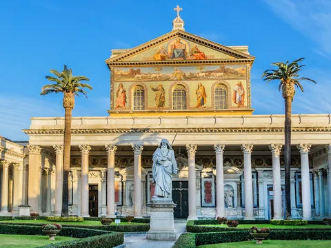 Fachada de la Basílica de San Pedro Extramuros en Roma, con columnas clásicas, mosaicos en el frontón y jardines con palmeras bajo cielo despejado.