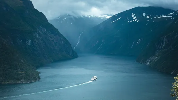 Barco navegando por el fiordo de Geiranger, Noruega, rodeado de montañas nevadas y cascadas bajo cielo nublado.