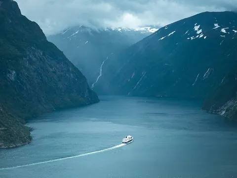 Barco navegando por el fiordo de Geiranger, Noruega, rodeado de montañas nevadas y cascadas bajo cielo nublado.