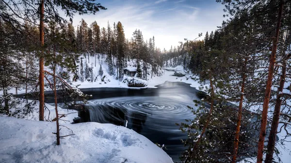Río parcialmente congelado con remolinos en el agua, rodeado de bosque nevado y una cabaña de madera, bajo un cielo azul claro."