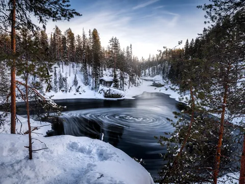 Río parcialmente congelado con remolinos en el agua, rodeado de bosque nevado y una cabaña de madera, bajo un cielo azul claro."