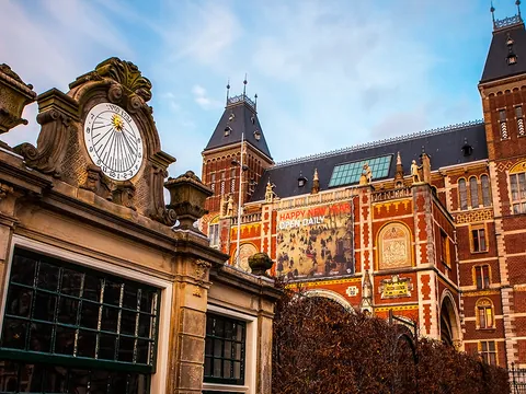 Fachada del Rijksmuseum en Ámsterdam al atardecer, con su arquitectura neorrenacentista de ladrillo rojo y una estructura de reloj solar ornamental en primer plano.