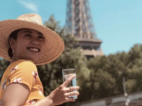 Mujer sonriente con sombrero y bebida frente a la Torre Eiffel en un día soleado.
