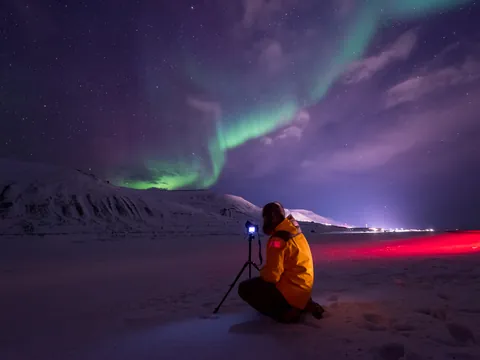 Persona con chaqueta amarilla fotografiando auroras boreales verdes sobre montañas nevadas, bajo un cielo estrellado con tonos púrpura.