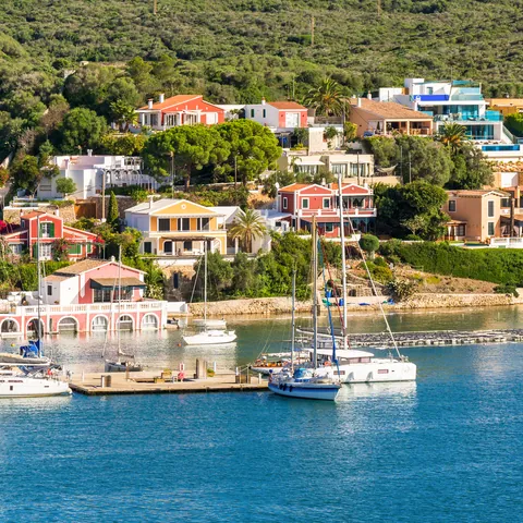 Veleros en el puerto de Mahón con casas y colinas de fondo.