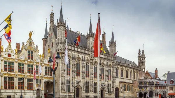 Plaza Burg en Brujas, Bélgica, con el Ayuntamiento gótico y edificios históricos adornados con banderas, bajo un cielo nublado.