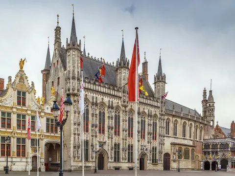 Plaza Burg en Brujas, Bélgica, con el Ayuntamiento gótico y edificios históricos adornados con banderas, bajo un cielo nublado.
