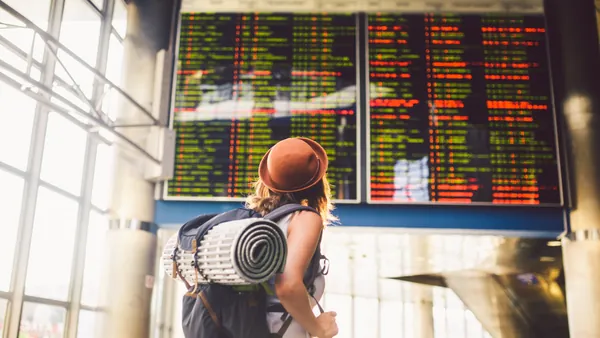 Chica con mochila y sombrero mirando el panel de información de vuelos en un aeropuerto, con listas de salidas y llegadas iluminadas en pantalla.