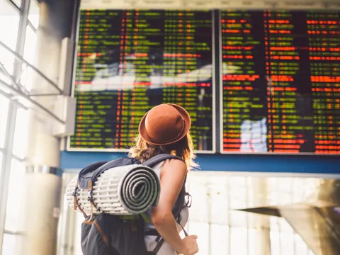 Chica con mochila y sombrero mirando el panel de información de vuelos en un aeropuerto, con listas de salidas y llegadas iluminadas en pantalla.