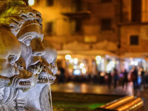 Primer plano de escultura en fuente romana con plaza iluminada al fondo de noche.