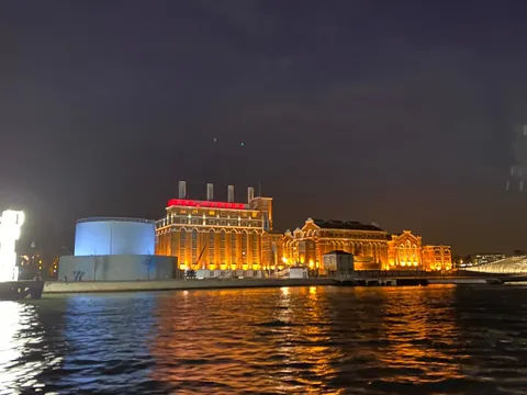 Vista nocturna de edificios históricos iluminados en la ribera del río Tajo en Lisboa, con reflejos de luces en el agua y cielo oscuro.