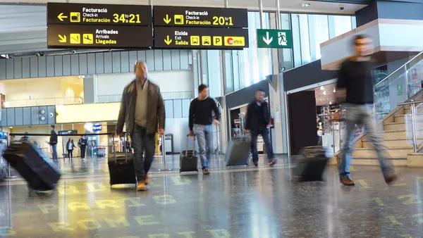 Interior de un aeropuerto con señalización de facturación y salidas, personas caminando con maletas de viaje.