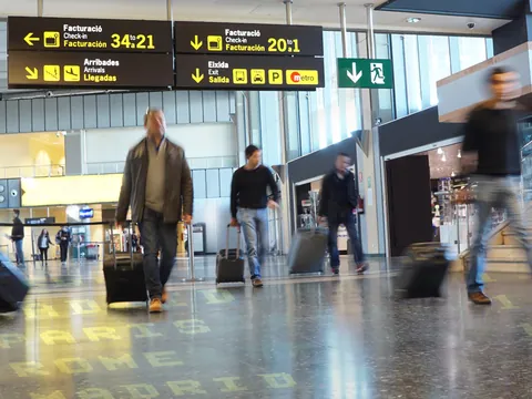 Interior de un aeropuerto con señalización de facturación y salidas, personas caminando con maletas de viaje.