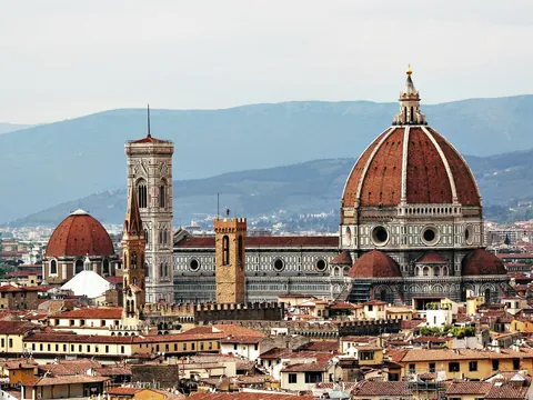 Vista panorámica de la Catedral de Santa María del Fiore en Florencia, Italia, destacando la cúpula de Brunelleschi, el campanario de Giotto y los tejados históricos de la ciudad con montañas al fondo.
