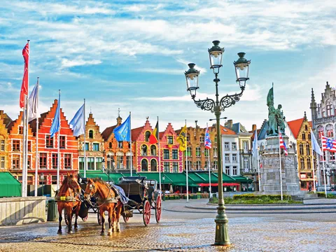 Plaza Mayor de Brujas con edificios flamencos de colores, banderas, farola antigua y carruaje tirado por caballos, bajo un cielo parcialmente nublado.