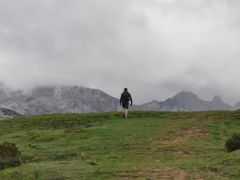 Senderista caminando por pradera en Lagos de Covadonga, Asturias, con montañas cubiertas de niebla.