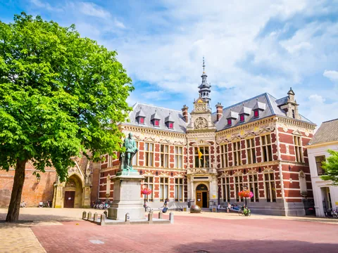 Edificio histórico de la Universidad de Utrecht en los Países Bajos, situado en una plaza abierta con árboles y una estatua en el centro.