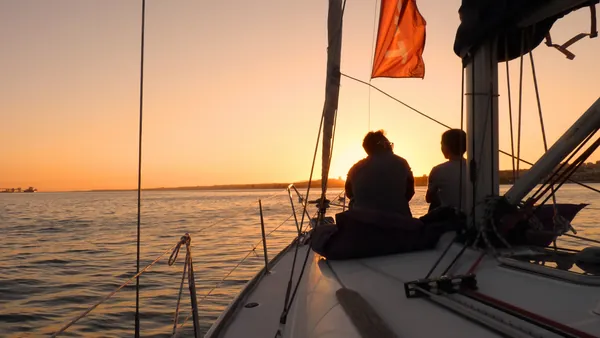 Dos personas sentadas en la proa de un velero durante la puesta de sol sobre el río Tajo en Lisboa, con el cielo iluminado en tonos cálidos.