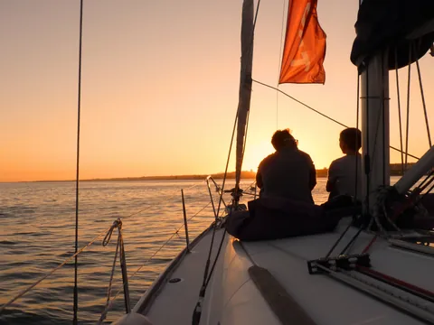 Dos personas sentadas en la proa de un velero durante la puesta de sol sobre el río Tajo en Lisboa, con el cielo iluminado en tonos cálidos.