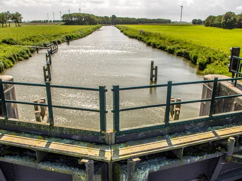 Compuerta y canal de agua en medio de campos verdes, con árboles y turbinas eólicas al fondo bajo un cielo nublado en un paisaje rural neerlandés.