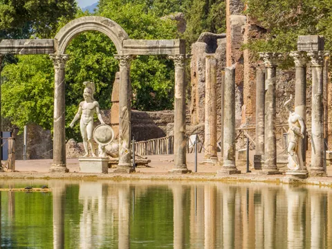 Estatuas y columnas romanas reflejadas en estanque rodeado de ruinas y vegetación en Tívoli.