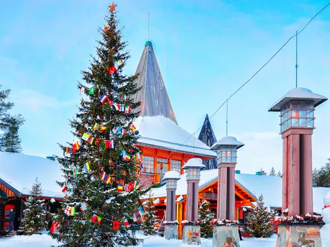 Árbol de Navidad decorado con banderas internacionales frente a una construcción de madera con techo nevado en la aldea de Santa Claus, Laponia, Finlandia.