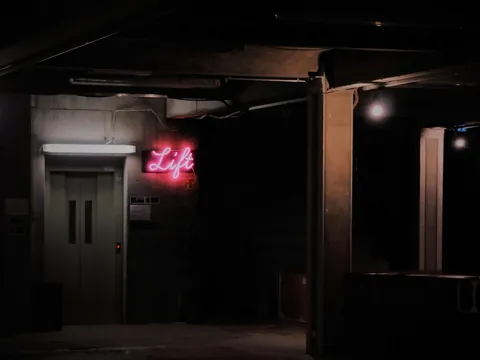 Entrada oscura con un cartel de neón rosa que indica “Lift”, en un ambiente industrial y alternativo
