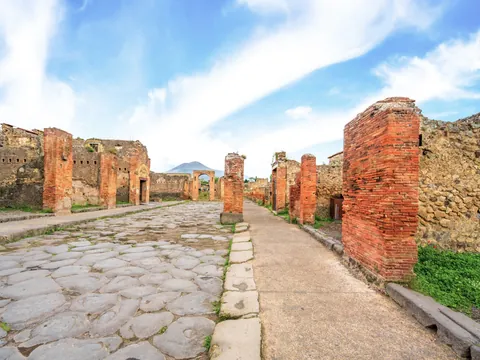 Calle romana empedrada en Pompeya con columnas y el Vesubio al fondo.