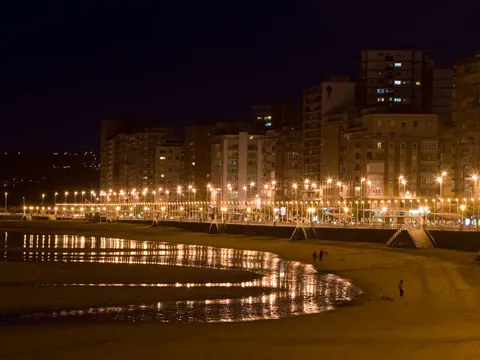 Vista nocturna del paseo marítimo y la playa de San Lorenzo en Gijón, con luces reflejadas sobre la arena mojada