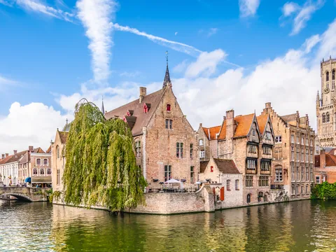 Canal de Brujas en Bélgica con edificios medievales de ladrillo y techos inclinados, terraza junto al agua y un árbol frondoso, bajo un cielo azul con nubes.