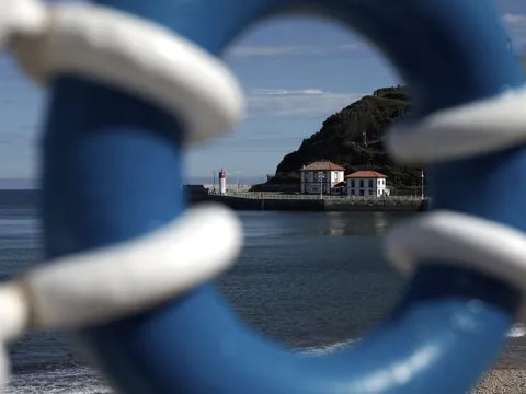 Vista del puerto de Ribadesella en Asturias, enmarcada por una barandilla azul con el faro y el mar Cantábrico al fondo.