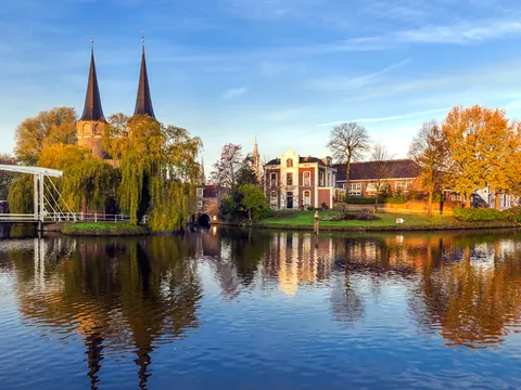 Vista de la Puerta del Este (Oostpoort) en Delft, Países Bajos, con dos altas torres góticas, árboles otoñales y edificios históricos reflejados en el agua tranquila de un canal.